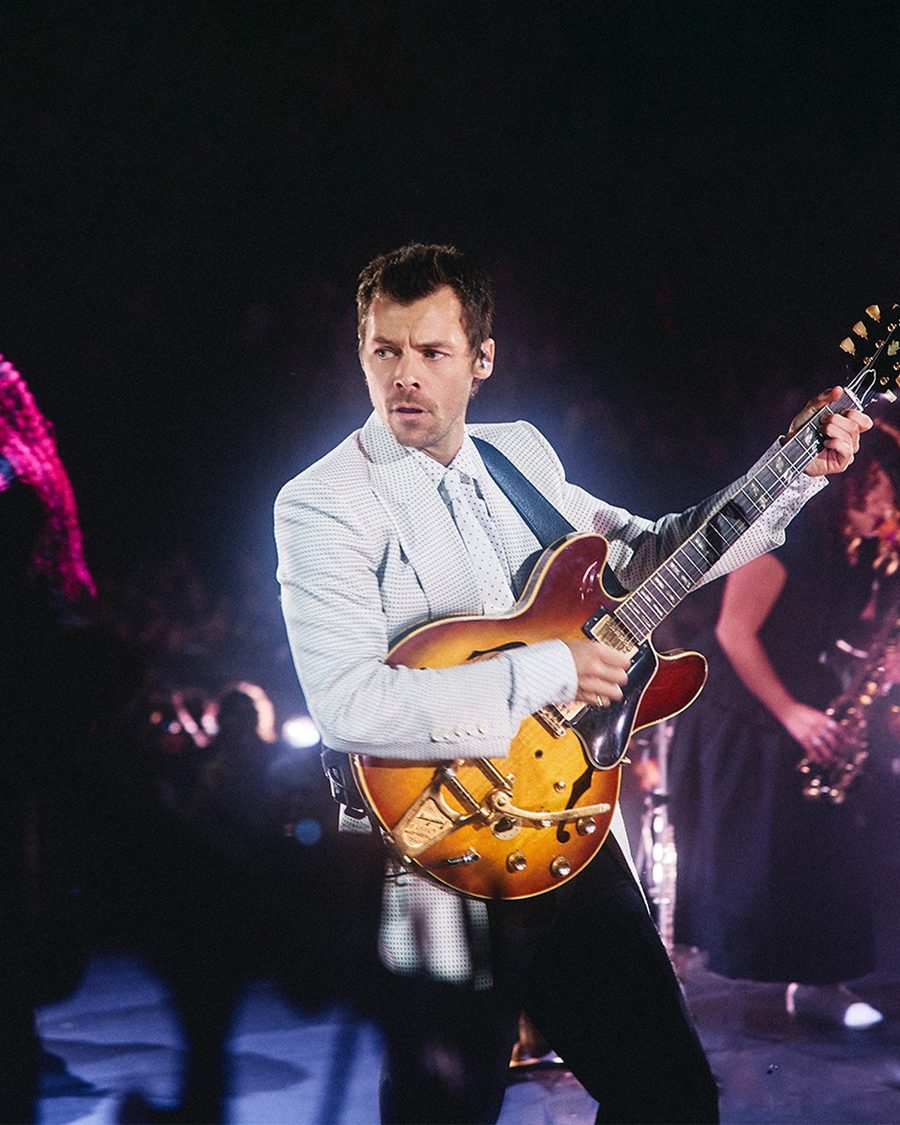 Harry Styles in a white suit playing a sunburst electric guitar on stage, band behind him under purple light